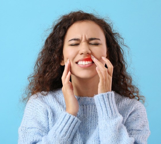 Woman in blue sweater touching her inflamed gums with eyes closed in pain