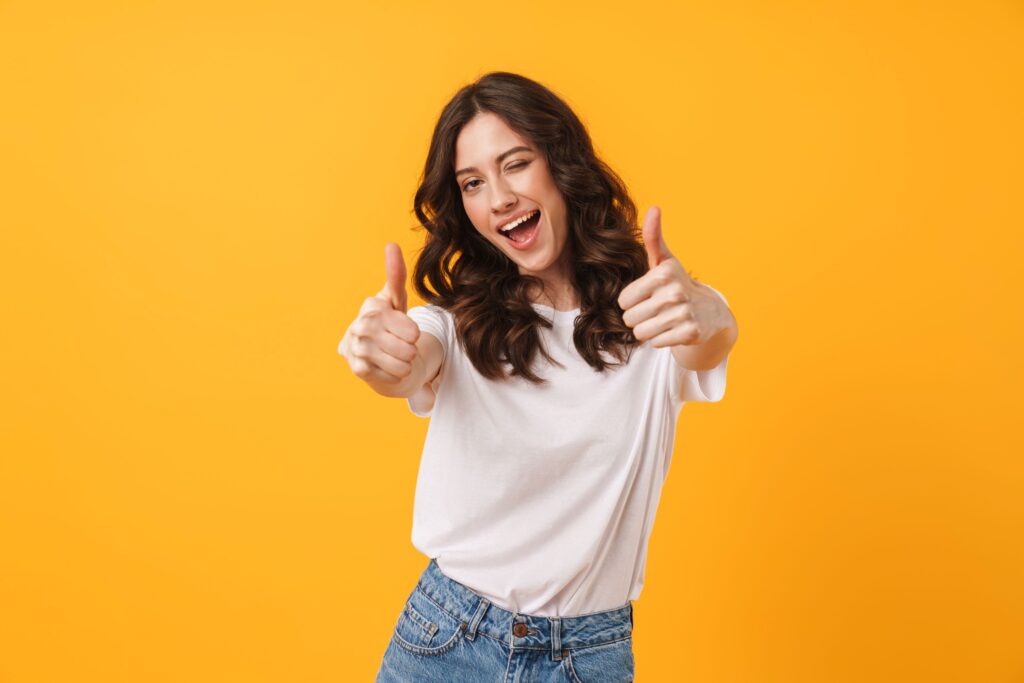 Woman with brown hair in white shirt smiling giving two thumbs up with yellow background